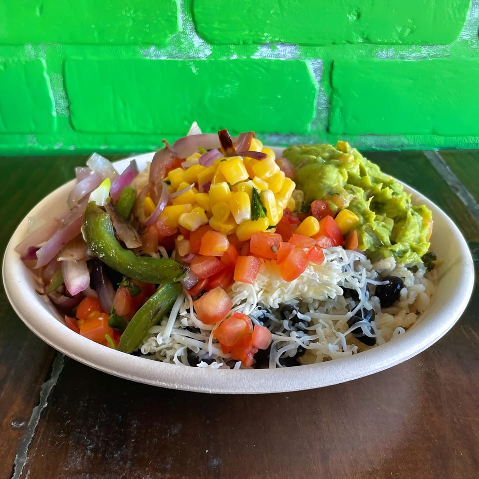 Colorful burrito bowl with rice, black beans, cheese, corn, tomatoes, guacamole, and grilled vegetables.